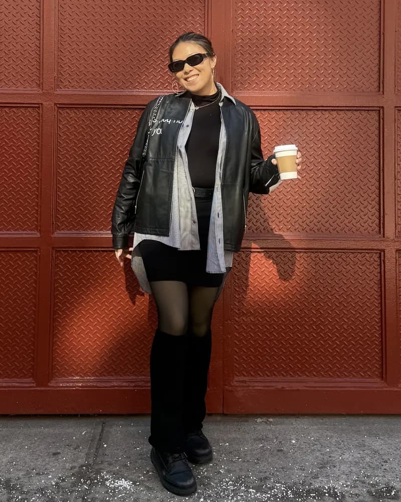 Charmaine in sunglasses, a black leather jacket and grey shirt, holding a coffee cup in front of a red paneled wall